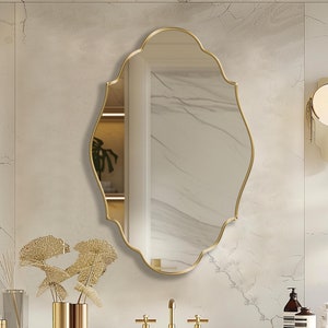 May include: A gold-framed oval mirror with a decorative, scalloped edge hangs above a white countertop with a gold faucet and two gold handles. The mirror is reflecting a white tiled wall with a gold light fixture.