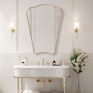 Puede incluir: Un tocador de baño blanco con un marco de metal dorado y un espejo dorado encima. El tocador tiene una encimera blanca con un grifo dorado y dos dispensadores de jabón dorados. Hay una toalla blanca en el estante inferior del tocador. Un jarrón con flores blancas está a la derecha del tocador.