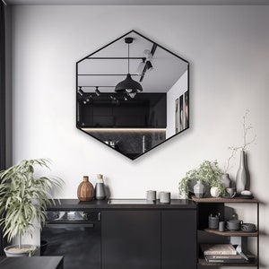 May include: A black hexagonal mirror with a beveled edge hangs on a white wall above a black countertop with a black oven and cabinets. The mirror reflects a kitchen with black cabinets and a black pendant light.