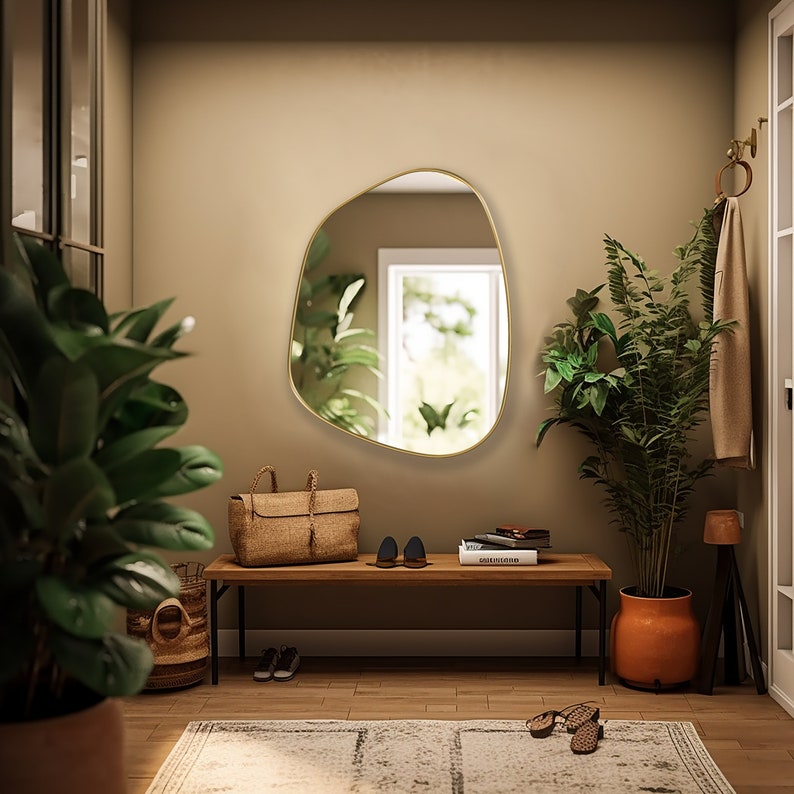 May include: A gold-framed, organic-shaped mirror hangs on a beige wall above a wooden bench. A potted plant with green leaves sits on the floor next to the bench. The bench has a woven bag and a pair of shoes on top.