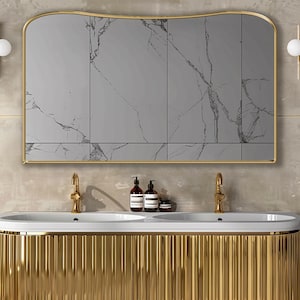 以下が含まれることがあります： 金色の仕上げが施された豪華なバスルームの洗面化粧台。金色のフレームの大きな鏡が特徴です。洗面化粧台には、金色の蛇口が付いたダブルシンクがあり、グローブライト付きの金色の燭台が両側にあります。鏡は大理石のような模様です。