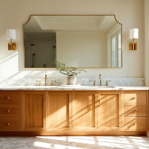 Könnte beinhalten: Ein helles Holzwaschbecken mit Marmorplatte und einem großen, goldgerahmten Spiegel. Der Waschtisch hat mehrere Schubladen und Schranktüren. Zwei goldene Wandleuchten flankieren den Spiegel. Der Raum hat eine neutrale Farbpalette.