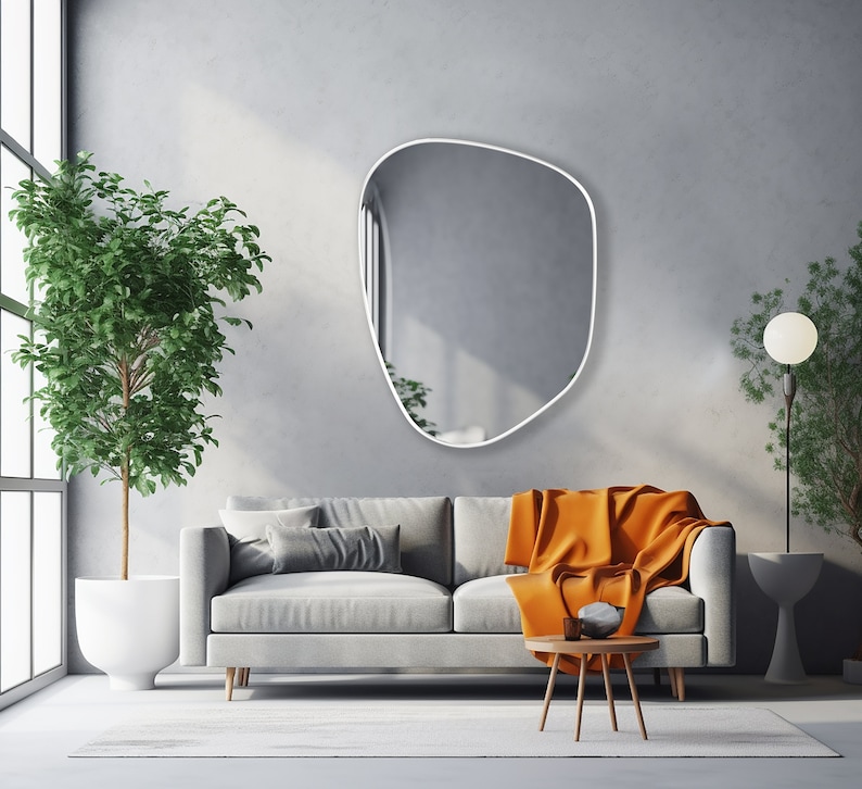 Asymmetrical mirror with white frame hung on the wall of a spacious living room over a big gray couch.