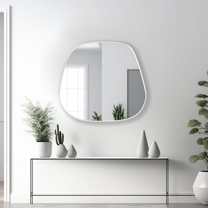 May include: A white, modern, free-form shaped wall mirror with a thin white frame hangs above a white console table with a black metal frame. The table is decorated with three small, gray, ceramic decorative objects and two potted plants. One plant is a tall, green, leafy plant in a white pot. The other plant is a small cactus in a white pot.