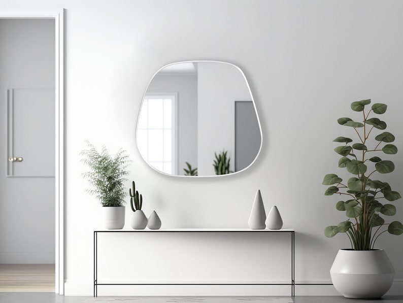 May include: A white, modern, free-form shaped wall mirror with a thin white frame hangs above a white console table with a black metal frame. The table is decorated with three small, gray, ceramic decorative objects and two potted plants. One plant is a tall, green, leafy plant in a white pot. The other plant is a small cactus in a white pot.
