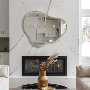 May include: A gold-framed, abstract-shaped mirror hangs above a black fireplace in a modern living room. The mirror reflects a white kitchen with a gold light fixture.