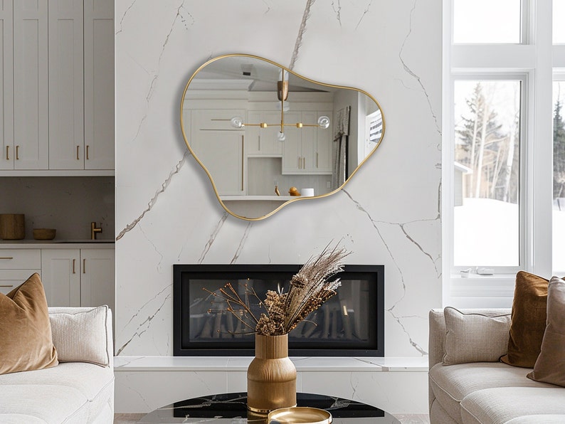 May include: A gold-framed, abstract-shaped mirror hangs above a black fireplace in a modern living room. The mirror reflects a white kitchen with a gold light fixture.