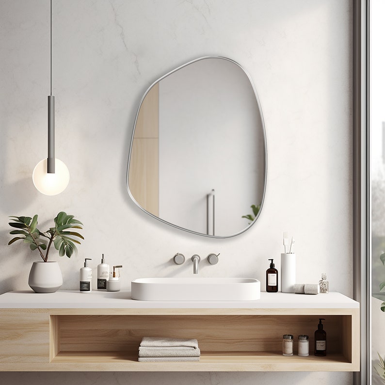 Asymmetrical mirror with silver frame hung on the wall of a bathroom over a washbasin.