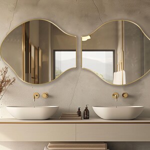 May include: Two white porcelain sinks with gold faucets and a gold-framed, wavy mirror above them. The mirror is split into two sections, each with a different view. The sinks are on a white countertop with a light brown backsplash.