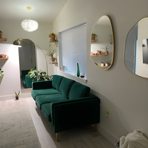 Two gold framed asymmetrical mirrors hung on the wall of a living room next to emerald color sofa in a spacious living room.