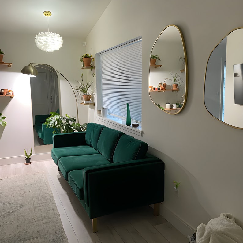 Two gold framed asymmetrical mirrors hung on the wall of a living room next to emerald color sofa in a spacious living room.