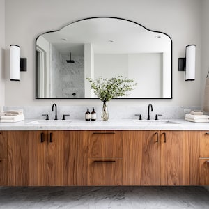 May include: A contemporary bathroom vanity with a large, arched mirror. The vanity has a wood grain cabinet with black hardware, a marble countertop, and two sinks with black taps. Two wall lights flank the mirror, and towels hang on the wall.