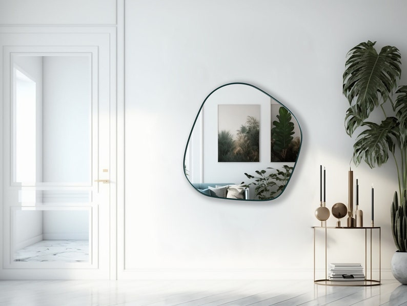 May include: A teal framed, freeform mirror hangs on a white wall above a gold metal console table with decorative objects. The mirror reflects two framed prints of nature scenes.