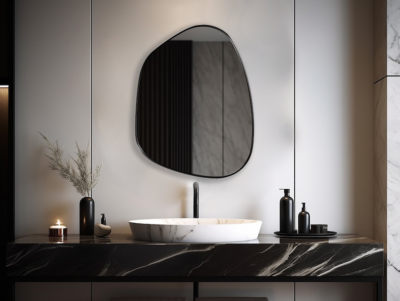 Asymmetrical mirror with black frame hung on the wall of a bathroom over a granite washbasin.