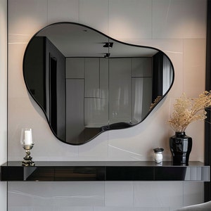 Könnte beinhalten: Ein schwarzer, unregelmäßig geformter Spiegel hängt über einem schwarzen Regal mit einem Kerzenhalter und einer Vase mit getrockneten Blumen. Der Spiegel spiegelt einen Raum mit weißen Wänden und einer schwarzen Tür wider.