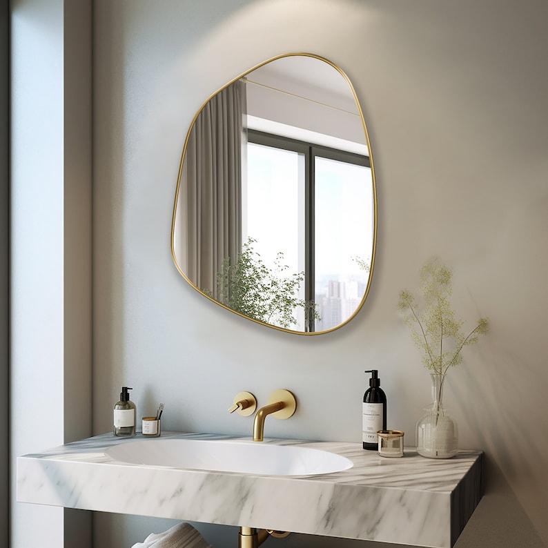 Asymmetrical mirror with gold frame hung on the wall of a bathroom over a white granite wash basin.