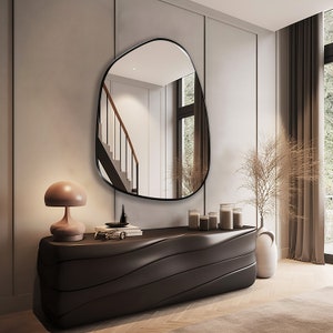 May include: A black, organic-shaped mirror hangs above a dark brown, wavy console table. A small, mushroom-shaped lamp sits on the table, along with a few decorative items. The mirror reflects a staircase with a wooden handrail.