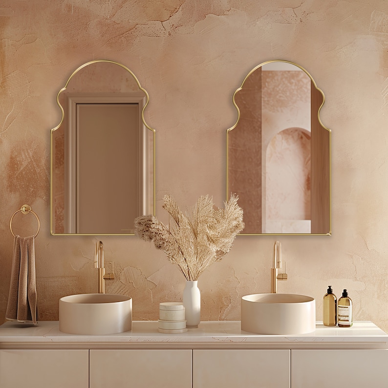 Two Bohemian Arched Mirrors with gold frame finish hung on the wall of a bathroom over a bathroom.