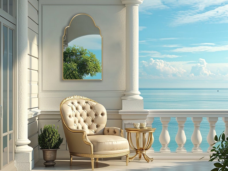 Arched Moroccan Mirror with gold frame finish hung on the wall of a terrace next to the sea, over an armchair.