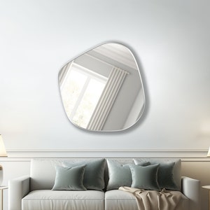 May include: A white couch with two green throw pillows and a beige throw blanket in front of a white wall with a large, irregular-shaped mirror hanging above it. Two lamps with white shades are on end tables on either side of the couch.