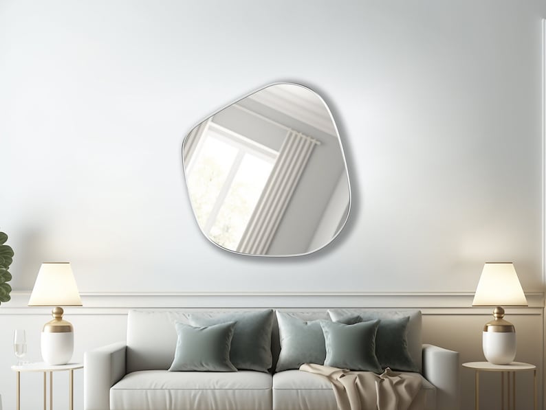 May include: A white couch with two green throw pillows and a beige throw blanket in front of a white wall with a large, irregular-shaped mirror hanging above it. Two lamps with white shades are on end tables on either side of the couch.