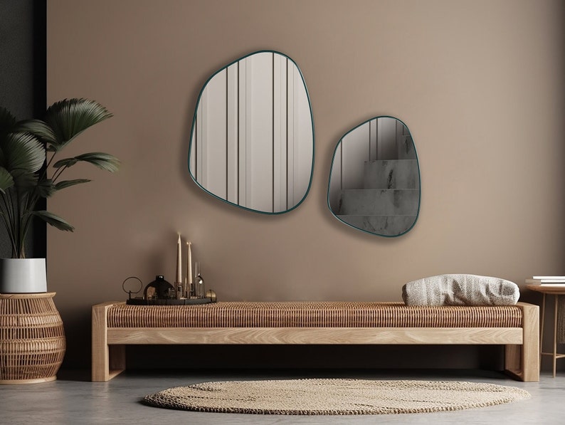 May include: Two organic shaped mirrors with teal frames hang on a light brown wall above a wooden bench. The mirrors reflect a staircase and a room with vertical blinds.