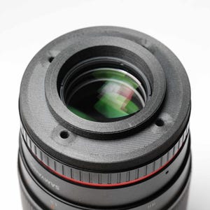 May include: Close-up of a black camera lens with a gray ring attachment. The lens has a red accent and the brand name "Samyang" is visible. The lens reflects green and other colors.
