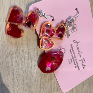 Op de afbeelding: Een paar rode hartvormige oorbellen met gouden accenten en glitter. De oorbellen liggen op een roze kaart met de tekst "Juwelen Fieke" en een websiteadres.
