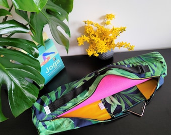 Peut inclure: Un sac de tapis de yoga noir avec un imprimé de feuilles tropicales. Le sac a une ouverture zippée et une sangle. Un tapis de yoga rose est à l'intérieur du sac. Une doublure en tissu jaune est visible à l'intérieur du sac. Un téléphone portable est dans la poche du sac. Le sac est sur une surface noire.