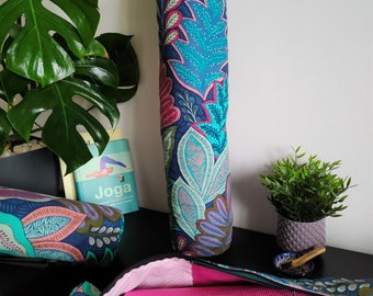 May include: A teal yoga mat bag with a floral pattern and a pink yoga mat inside. The bag has a black adjustable strap and a zipper closure. The bag is sitting on a black surface with a teal yoga mat and a book titled "Joga" on the surface.