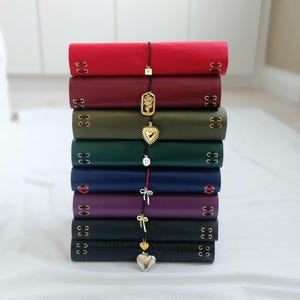 May include: A vertical stack of eight leather-bound journals in various colors including red, burgundy, olive green, dark green, navy blue, purple, and black. Each journal features side lacing with metal eyelets and a dangling charm on a cord.