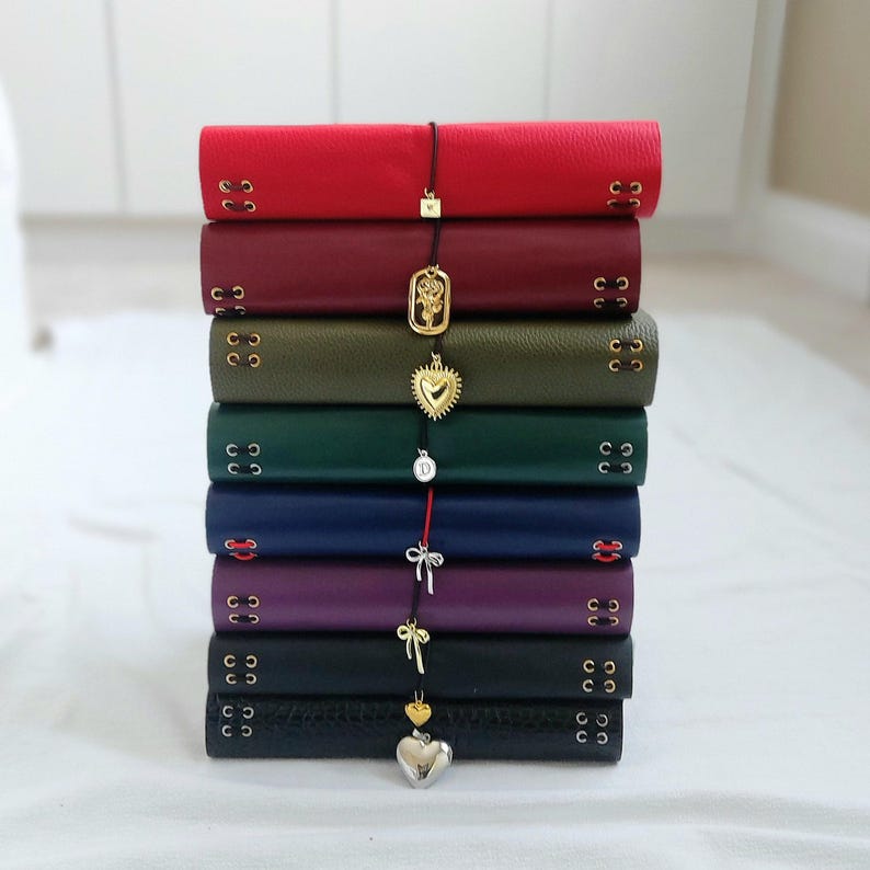May include: A vertical stack of eight leather-bound journals in various colors including red, burgundy, olive green, dark green, navy blue, purple, and black. Each journal features side lacing with metal eyelets and a dangling charm on a cord.