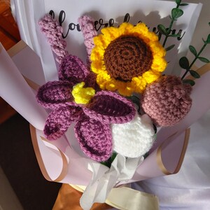 May include: A handmade crochet bouquet featuring a large sunflower with yellow petals and a brown center, a purple flower, and a white flower. The bouquet is wrapped in pink paper and tied with a white ribbon. The bouquet includes a sprig of green leaves.