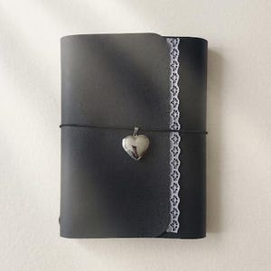 May include: A black journal with a smooth cover featuring a vertical strip of white lace trim along the right edge. A thin black elastic cord wraps horizontally around the center, holding a small, polished silver-toned heart-shaped locket charm.
