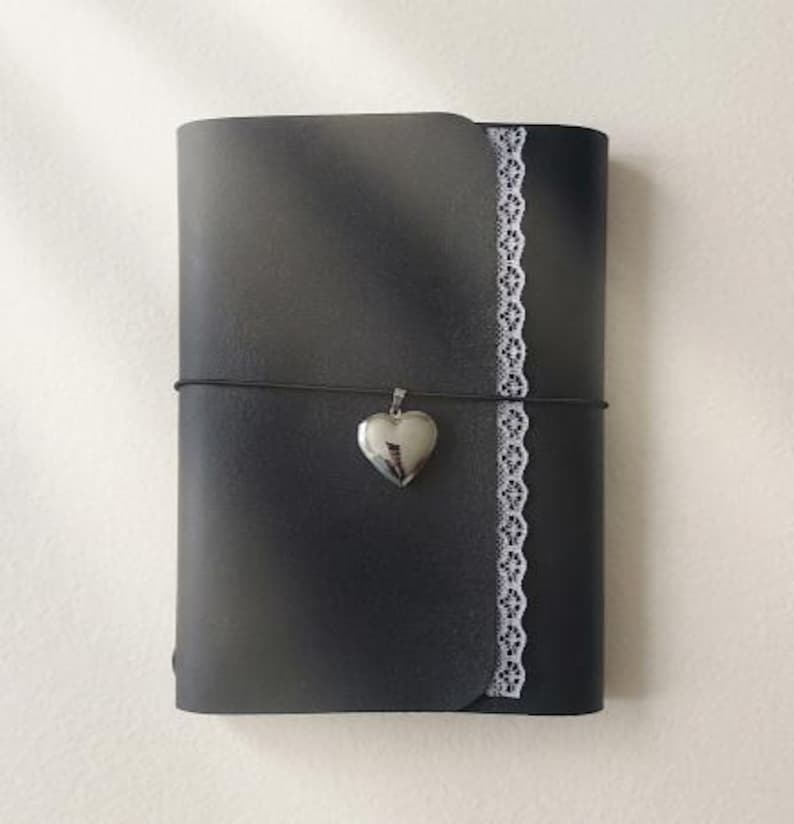 May include: A black journal with a smooth cover featuring a vertical strip of white lace trim along the right edge. A thin black elastic cord wraps horizontally around the center, holding a small, polished silver-toned heart-shaped locket charm.