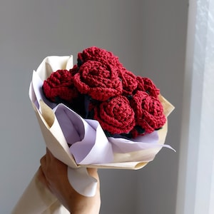 Puede incluir: Un ramo de rosas rojas hechas a mano y tejidas a ganchillo. Las flores est&aacute;n bien enrolladas y dispuestas en un manojo, envueltas en papel marr&oacute;n y blanco. El ramo se sostiene en una mano sobre un fondo neutro.