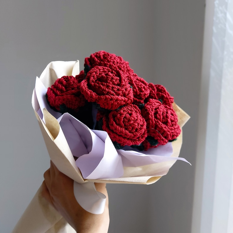 Puede incluir: Un ramo de rosas rojas hechas a mano y tejidas a ganchillo. Las flores est&aacute;n bien enrolladas y dispuestas en un manojo, envueltas en papel marr&oacute;n y blanco. El ramo se sostiene en una mano sobre un fondo neutro.