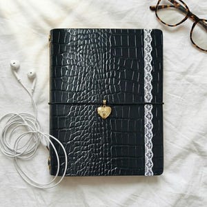 May include: A black notebook with a textured crocodile skin pattern, featuring a vertical white lace trim on the right side. A gold heart-shaped locket hangs from a black elastic band wrapped horizontally around the center of the notebook cover.