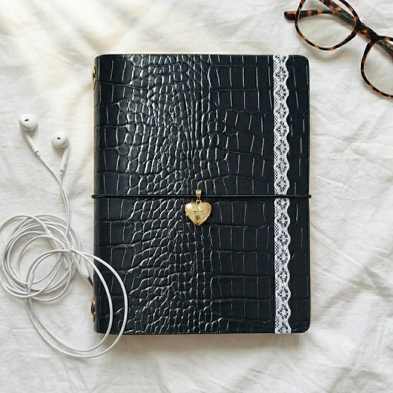 May include: A black notebook with a textured crocodile skin pattern, featuring a vertical white lace trim on the right side. A gold heart-shaped locket hangs from a black elastic band wrapped horizontally around the center of the notebook cover.