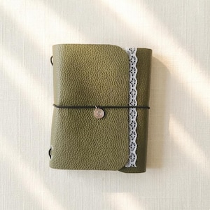 May include: An olive green journal with a textured pebble grain finish, featuring a vertical strip of white lace trim along the right edge. A thin black elastic band wraps around the center, secured by a small circular charm embossed with the letter S.