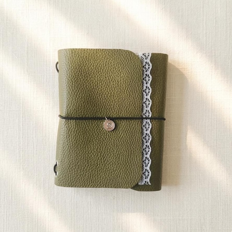 May include: An olive green journal with a textured pebble grain finish, featuring a vertical strip of white lace trim along the right edge. A thin black elastic band wraps around the center, secured by a small circular charm embossed with the letter S.