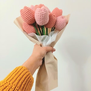 May include: A bouquet of handmade crochet tulips in varying shades of pink, wrapped in brown paper. The stems are green, and the flowers are held together with white fabric. The bouquet is a craft item.