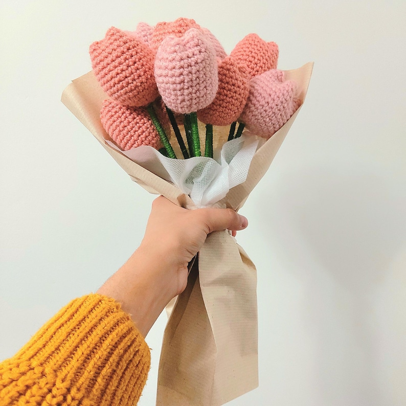 May include: A bouquet of handmade crochet tulips in varying shades of pink, wrapped in brown paper. The stems are green, and the flowers are held together with white fabric. The bouquet is a craft item.