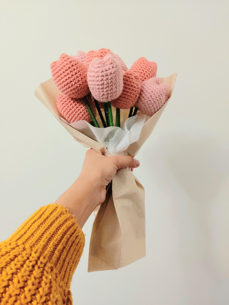 May include: A bouquet of handmade crochet tulips in shades of pink and peach. The flowers are wrapped in brown paper and tied with white tulle. The stems are green.