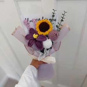 May include: A handmade crochet bouquet featuring a sunflower, purple flower, and other crocheted flowers. The bouquet is wrapped in lavender paper and tied with a cream ribbon. The bouquet includes a card with the text "I love you".