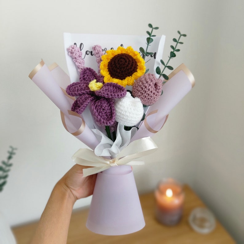 May include: A handmade crochet bouquet featuring a sunflower, purple iris, and other flowers. The bouquet is wrapped in lavender paper and tied with a cream ribbon. The flowers are in shades of yellow, purple, white, and pink.