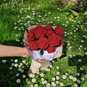 Puede incluir: Un ramo de rosas rojas de ganchillo, envuelto en papel marr&oacute;n y papel de seda blanco. Las flores se presentan sobre un fondo de hierba verde y peque&ntilde;as flores blancas.