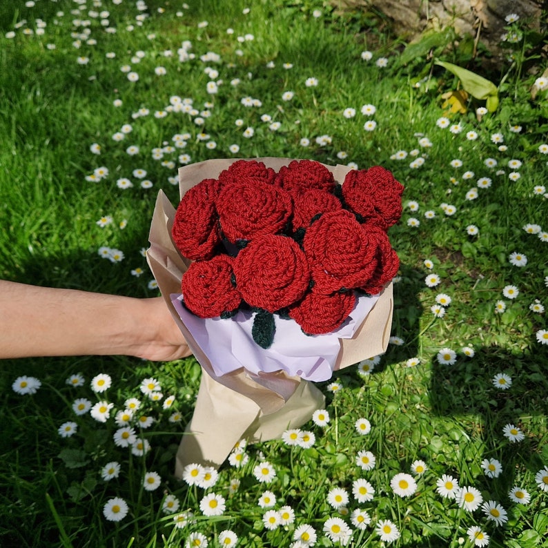 Puede incluir: Un ramo de rosas rojas de ganchillo, envuelto en papel marr&oacute;n y papel de seda blanco. Las flores se presentan sobre un fondo de hierba verde y peque&ntilde;as flores blancas.