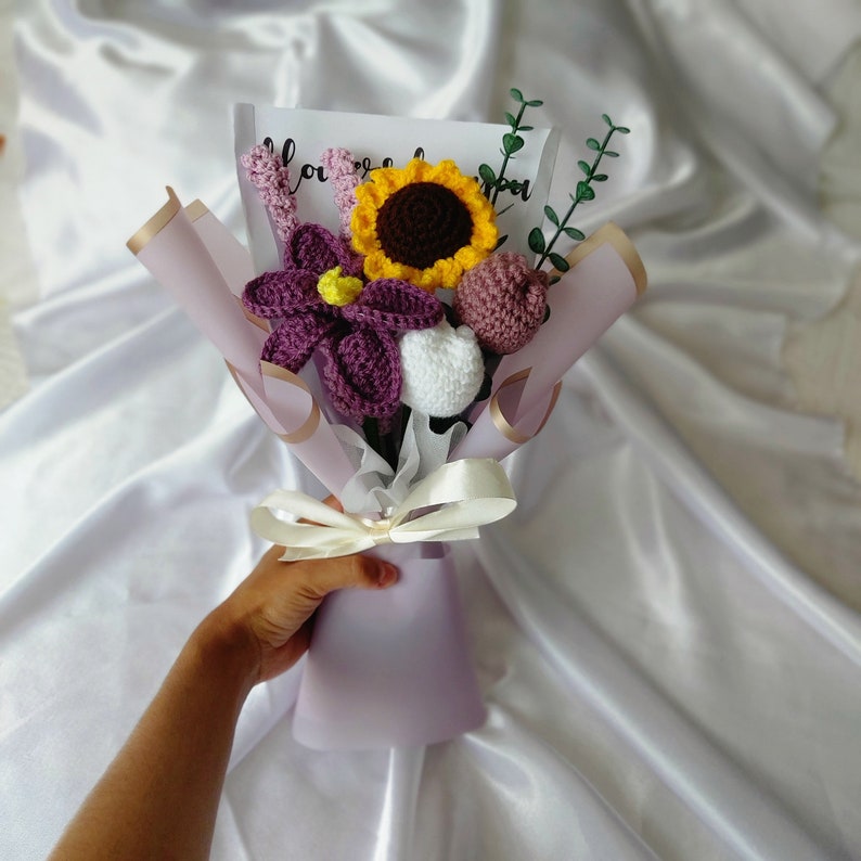 May include: A handmade crochet bouquet featuring a sunflower, purple flower, white flower, and other floral elements. The bouquet is wrapped in lavender paper and tied with a cream ribbon. A card with text is visible.