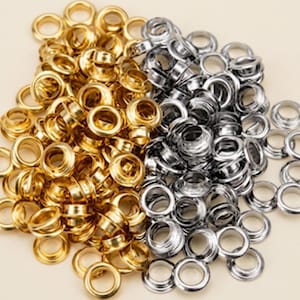 May include: A large collection of metal eyelets in two distinct finishes, featuring a shiny gold-tone color on the left side and a polished silver-tone color on the right side. These circular hardware components have a hollow center and a rim.